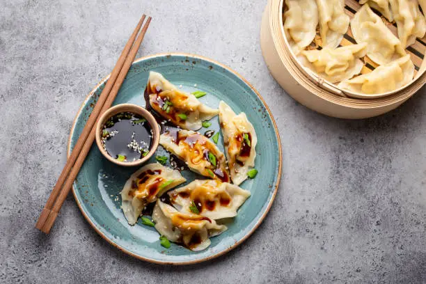 Close-up, top view of traditional Asian/Chinese dumplings in blue plate with soy sauce, chopsticks and a bamboo steamer on gray rustic stone background. Authentic Chinese cuisine