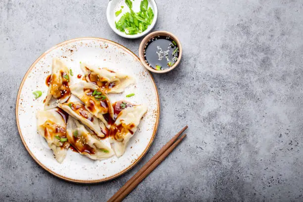 Close-up, top view of traditional Asian/Chinese dumplings on white plate with soy sauce and chopsticks on gray rustic stone background with space for text. Authentic Chinese cuisine, copy space