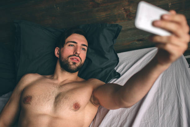 naked bearded dark-hair handsome man shirtless in white bed taking a selfie - sensuality lifestyles cheerful comfortable imagens e fotografias de stock