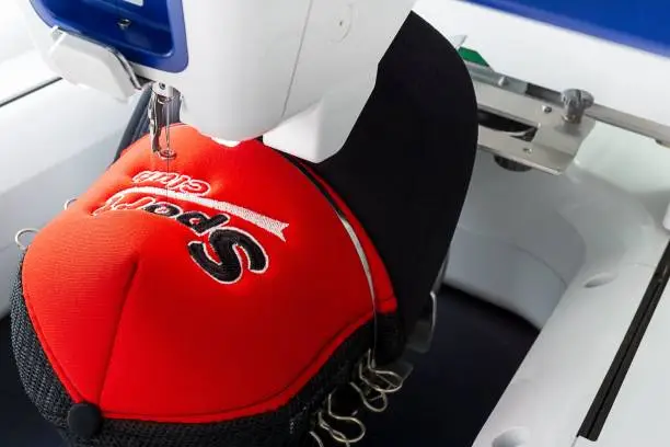 Close up image of white embroidery machine embroidering logo on red and black sport cap