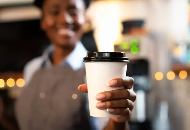 A smiling barista holding out a takeaway coffee towards the camera. Selective focus, with focus on the disposable cup.
