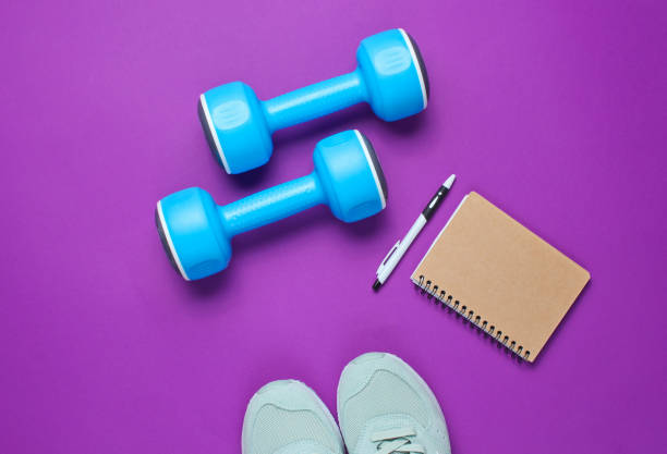 istock Fitness concept, workout plan. Sport shoes, plastic blue dumbbells, notepad on purple background. Top view. Flat lay 1138884532