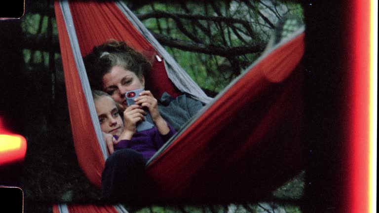 Retro style film footage of mother and daughter with smartphone sharing a cozy hammock on family camping trip to Rio Grande del Norte National Monument.