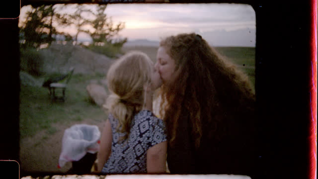 Retro style film footage of mother and daughter sharing a sweet kiss at picnic table and daughter turning to kiss camera on family camping trip.