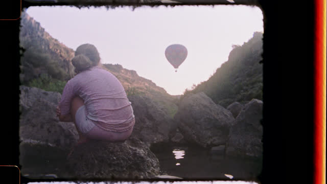 Nostalgic film footage of daydreaming young girl looking up at hot air balloon from rocky perch at Black Rock Hot Springs.