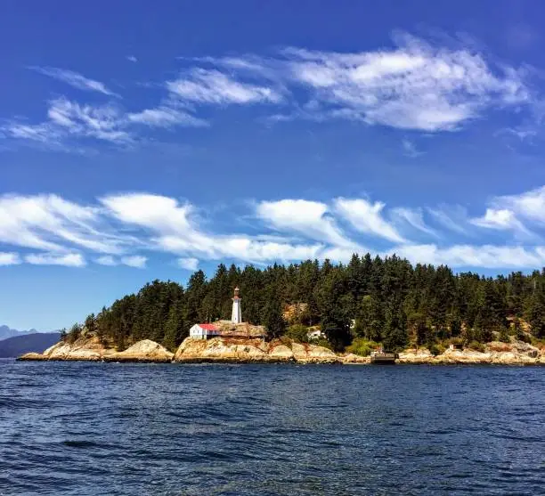 Photo of A far away view of the Point Atkinson Lighthouse in West Vancouver, British Columbia, Canada.  The lighthouse is red and white and sits along a rocky point covered in evergreen forest.  It is sunny.