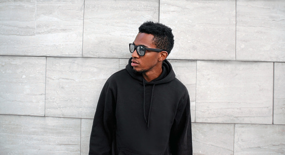 Portrait african man in black hoodie, sunglasses looking away on city street over gray brick wall background