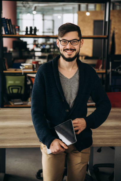 istock Handsome guy with a charismatic smile, with a beard and glasses, stylishly dressed, stands indoors with a tablet in his hands 1138939287