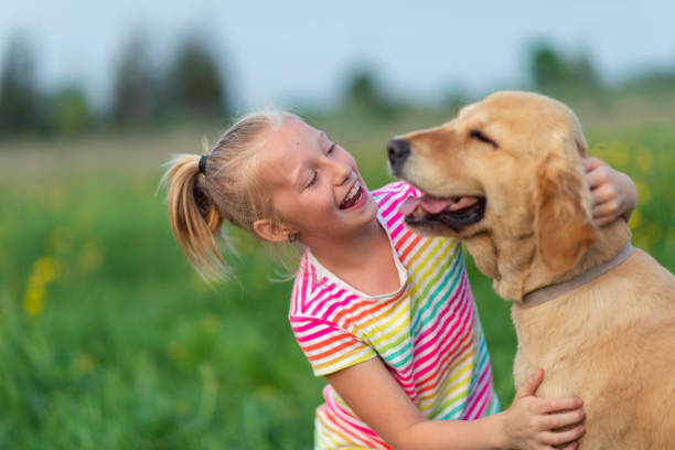 A young girl smiles as she pets her dog affectionately. She is sitting outside in the grass on a sunny day.