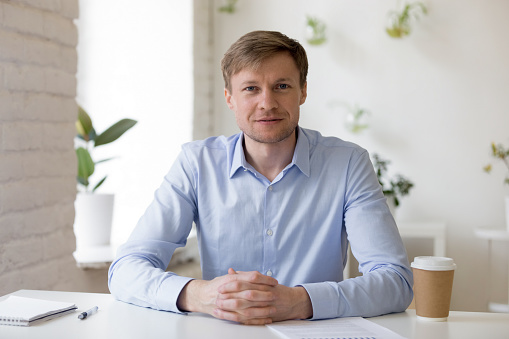 Smiling friendly millennial businessman sitting at workplace, looking at camera, confident successful ceo, happy young professional, business coach or executive manager head shot portrait