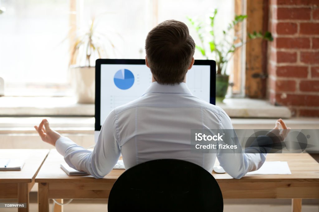 Rear view businessman sitting at desk opposite pc doing yoga Rear back view businessman sitting at desk opposite pc doing yoga feels placidity and calmness. Employee take break resting relaxing practicing meditation. Healthy lifestyle and stress relief concept Rear View Stock Photo