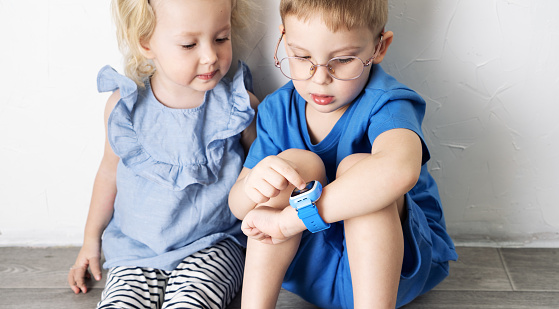 A little boy child in a blue T-shirt and glasses shows the girl his smart watch and presses a finger. Portrait against white wall.