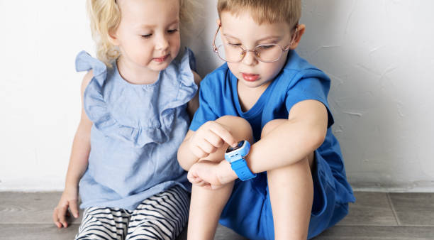 A little boy child in a blue T-shirt and glasses shows the girl his smart watch and presses a finger. A little boy child in a blue T-shirt and glasses shows the girl his smart watch and presses a finger. Portrait against white wall. blue brothers stock pictures, royalty-free photos & images