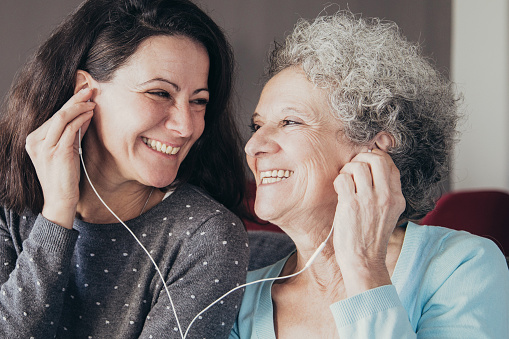 Happy senior woman and her daughter listening to music together. Mother and daughter wearing earphones and sitting on sofa with home interior in background. Happy senior mother and daughter concept.