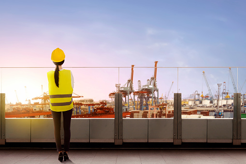 Rear view of asian female worker with safety vest and hard hat standing on office terrace and looking at dock with sunset view background