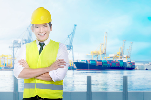 Handsome asian worker with yellow hard hat standing on the office with industrial harbor and blue sky background