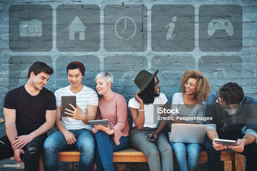 We stay connected everywhere we go Shot of a group of cheerful creative businesspeople using their digital devices while sitting on a bench together outdoors Gamification Stock Photo