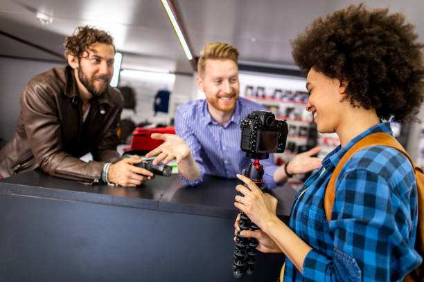 vendedor de loja apresentando os recursos de gravação de vídeo de uma câmera dslr para um jovem vlogger feminino e seu parceiro em uma loja de câmeras - retail occupation flash - fotografias e filmes do acervo