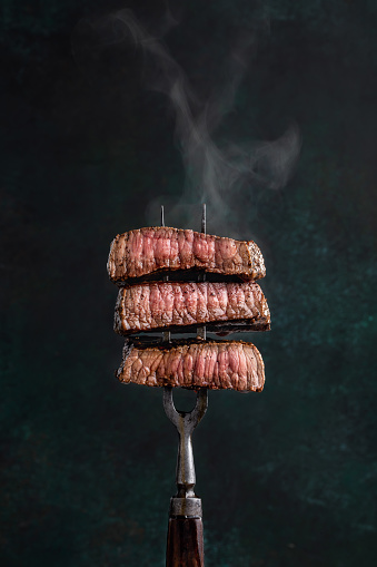 Slices of beef steak with steam on vintage fork on dark background