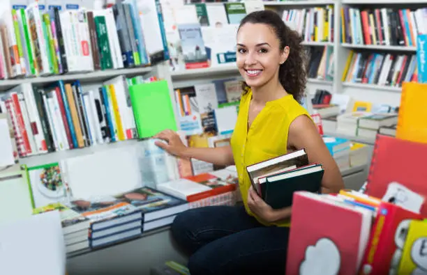 Young smiling female student holding books in hard cover in shop