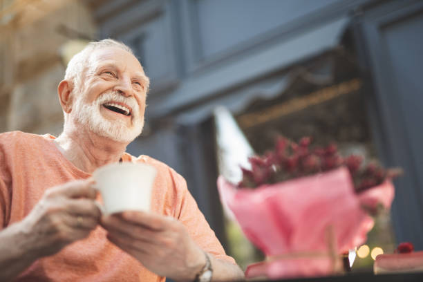Laughing elderly man drinking tea outdoors Low angle waist up portrait of delighted mature male sitting at table outside. He is holding cup of hot drink and looking with pleasure and content. Copy space in right side senior men stock pictures, royalty-free photos & images