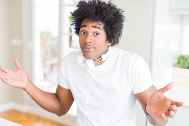african american man wearing headphones listening to music clueless and confused expression with arms and hands raised. doubt concept. - head and shoulders audio imagens e fotografias de stock
