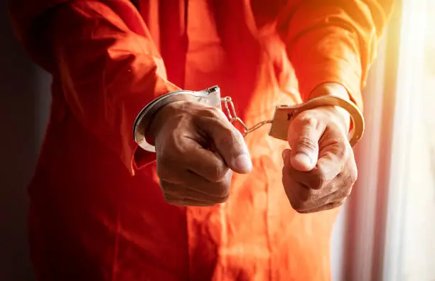 Photo of close up of prisoner's hands with handcuffs in orange jumpsuit at prison