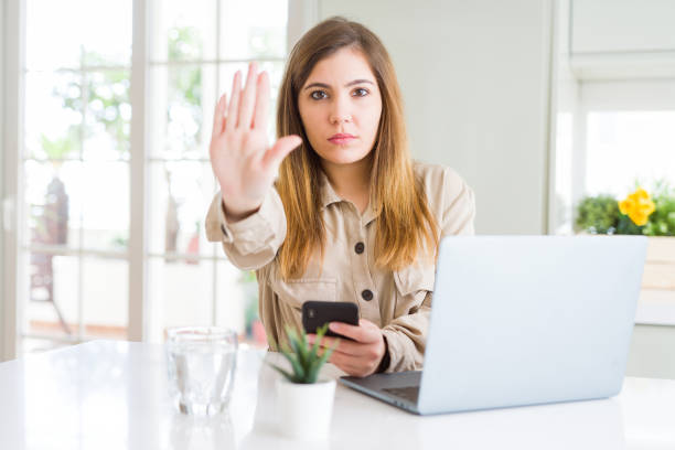 istock Beautiful young woman using smartphone and computer with open hand doing stop sign with serious and confident expression, defense gesture 1139053268