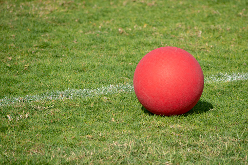 A red playground ball sits next to the white line on a green grass field.