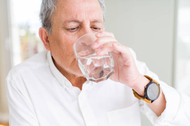 bell'uomo anziano che beve un bicchiere d'acqua fresco a casa - thirsty foto e immagini stock