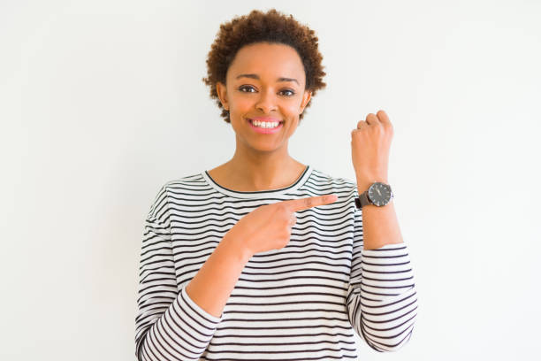 若い美しいアフリカ系アメリカ人女性のストライプのセーターを着て白の背景の上に急いで時間、焦り、動揺と怒っている締め切りの遅延を指しています - checking the time women impatient wristwatch ストックフォトと画像