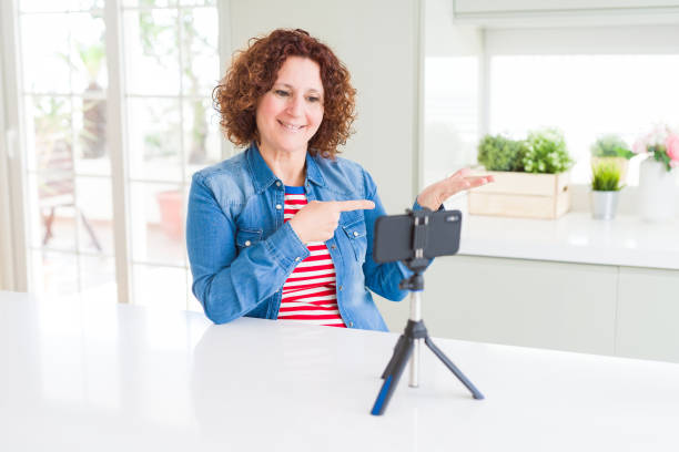 istock Senior woman doing video chat using smartphone camera amazed and smiling to the camera while presenting with hand and pointing with finger. 1139059773