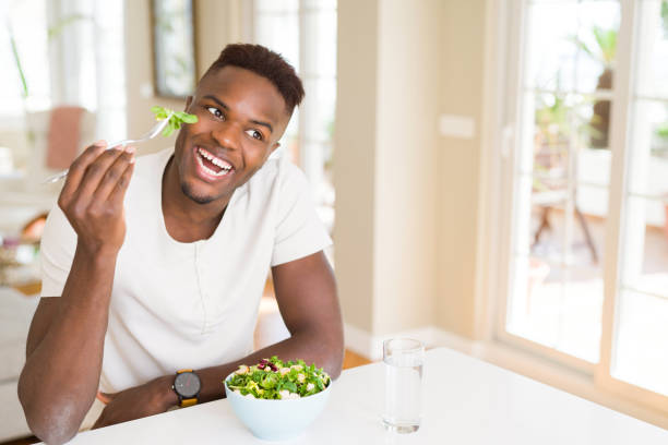 istock Handsome african young man eating a healthy vegetable salad using a fork to eat lettuce, happy and smiling sitting on the table 1139060230