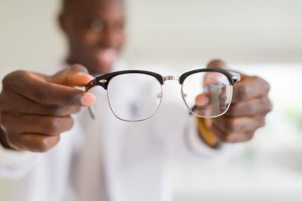 hombre óptico afroamericano que sostiene y muestra lentes de gafas a clientes en la tienda de óptica mientras sonríe confiado - human eye eyesight optometrist lens fotografías e imágenes de stock
