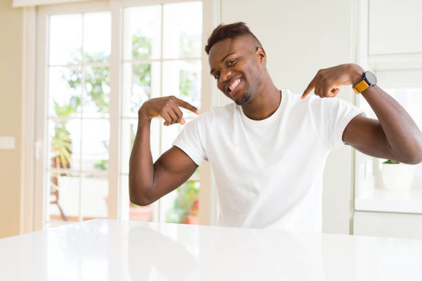 istock Handsome african american man on white table at home looking confident with smile on face, pointing oneself with fingers proud and happy. 1139060891