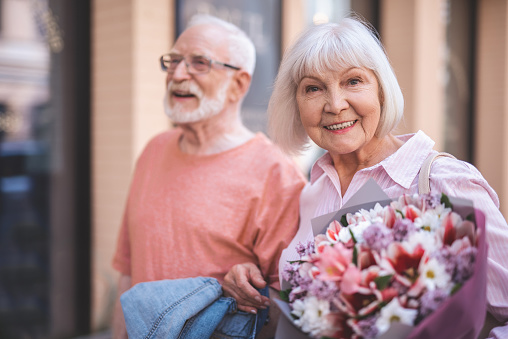 Waist up portrait of smiling senior lady strolling with male outside. She is happily holding bunch of flowers and looking at camera with joy
