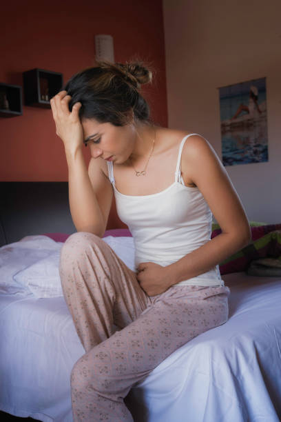 Hispanic cute young woman with stomachache or PMS pain while sitting on bed stock photo