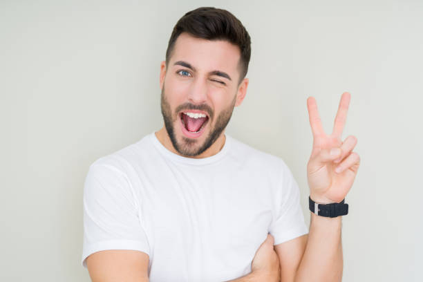 Young handsome man wearing casual white t-shirt over isolated background smiling with happy face winking at the camera doing victory sign. Number two. Young handsome man wearing casual white t-shirt over isolated background smiling with happy face winking at the camera doing victory sign. Number two. young man wink stock pictures, royalty-free photos & images