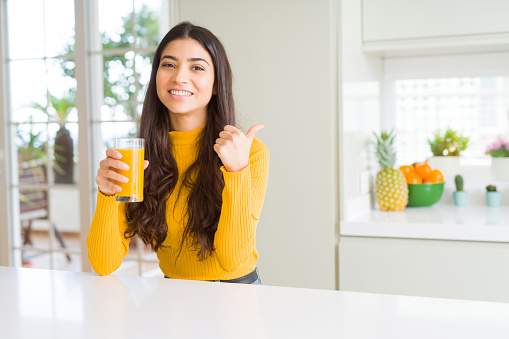 Young woman drinking a glass of fresh orange juice pointing and showing with thumb up to the side with happy face smiling
