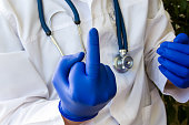 Fuck you sign fingers of hand by doctor or health worker. Doctor showing obscene sign fuck you with raised of middle finger of hand, dressed in blue latex glove in white medical gown on background