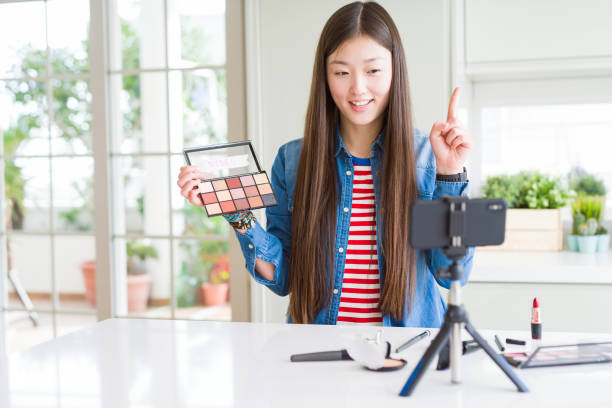 istock Beautiful Asian influencer woman recording make up tutorial using smartphone camera surprised with an idea or question pointing finger with happy face, number one 1139071185