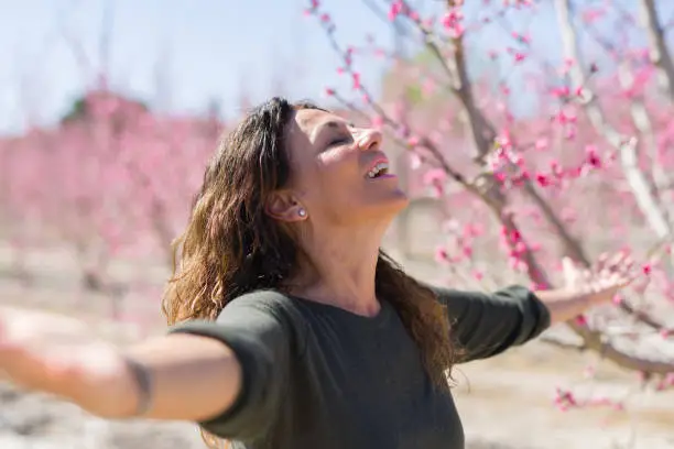 Beautiful middle age woman in the middle of pink peach flowers and trees smiling cheerful with open arms enjoying sunbathe on sunny day