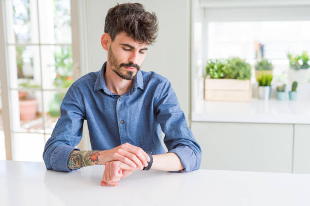 Young man wearing casual shirt sitting on white table Checking the time on wrist watch, relaxed and confident Young man wearing casual shirt sitting on white table Checking the time on wrist watch, relaxed and confident wrist tattoo stock pictures, royalty-free photos & images