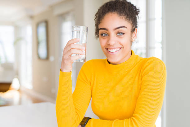 jeune femme afro-américaine buvant un verre d’eau fraîche avec un visage heureux debout et souriant avec un sourire confiant montrant des dents - water women glass healthy eating photos et images de collection