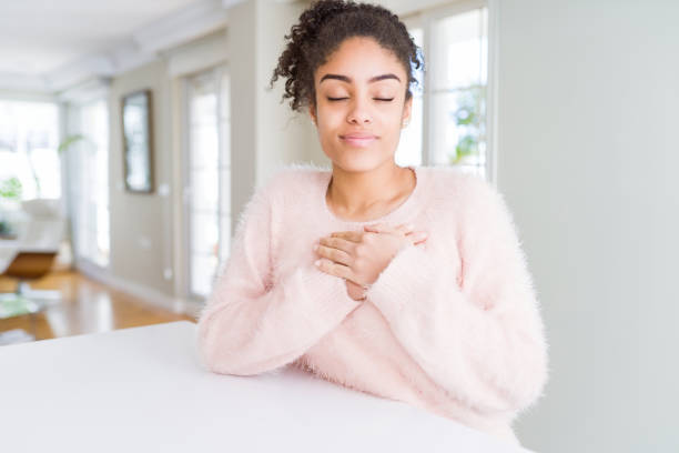 bella giovane donna afroamericana con i capelli afro sorridenti con le mani sul petto con gli occhi chiusi e gesto grato sul viso. concetto di salute. - male smiling african descent black foto e immagini stock