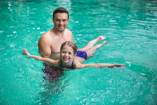 Waist up portrait of delighted man holding his daughter in water and teaching her floating. Small girl is lying on father hands with smile and content
