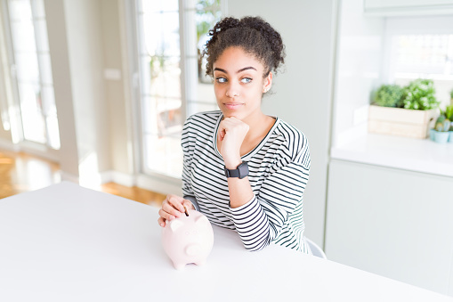 Young african american woman putting a coin inside piggy bank as invest or saving serious face thinking about question, very confused idea