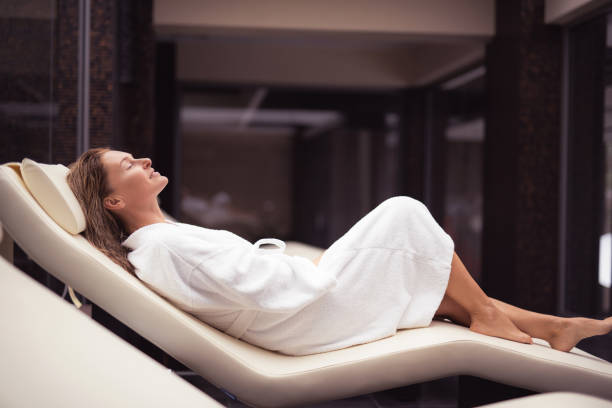 Beautiful middle aged woman enjoying rest at spa lounge Pure pleasure. Side view portrait of charming lady with closed eyes lying on daybed after shower. She is wearing soft white bathrobe and smiling chaise longue woman stock pictures, royalty-free photos & images