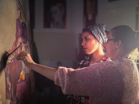 Young artist teaching young female Latino student how to painting, dramatic light