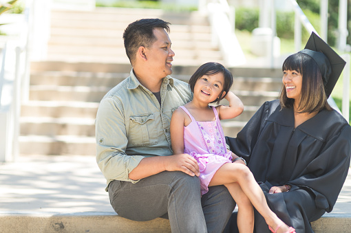 A middle-aged graduate wearing her cap and gown sits on campus steps with her husband and their young daughter. Mom and dad are looking affectionately at each other.
